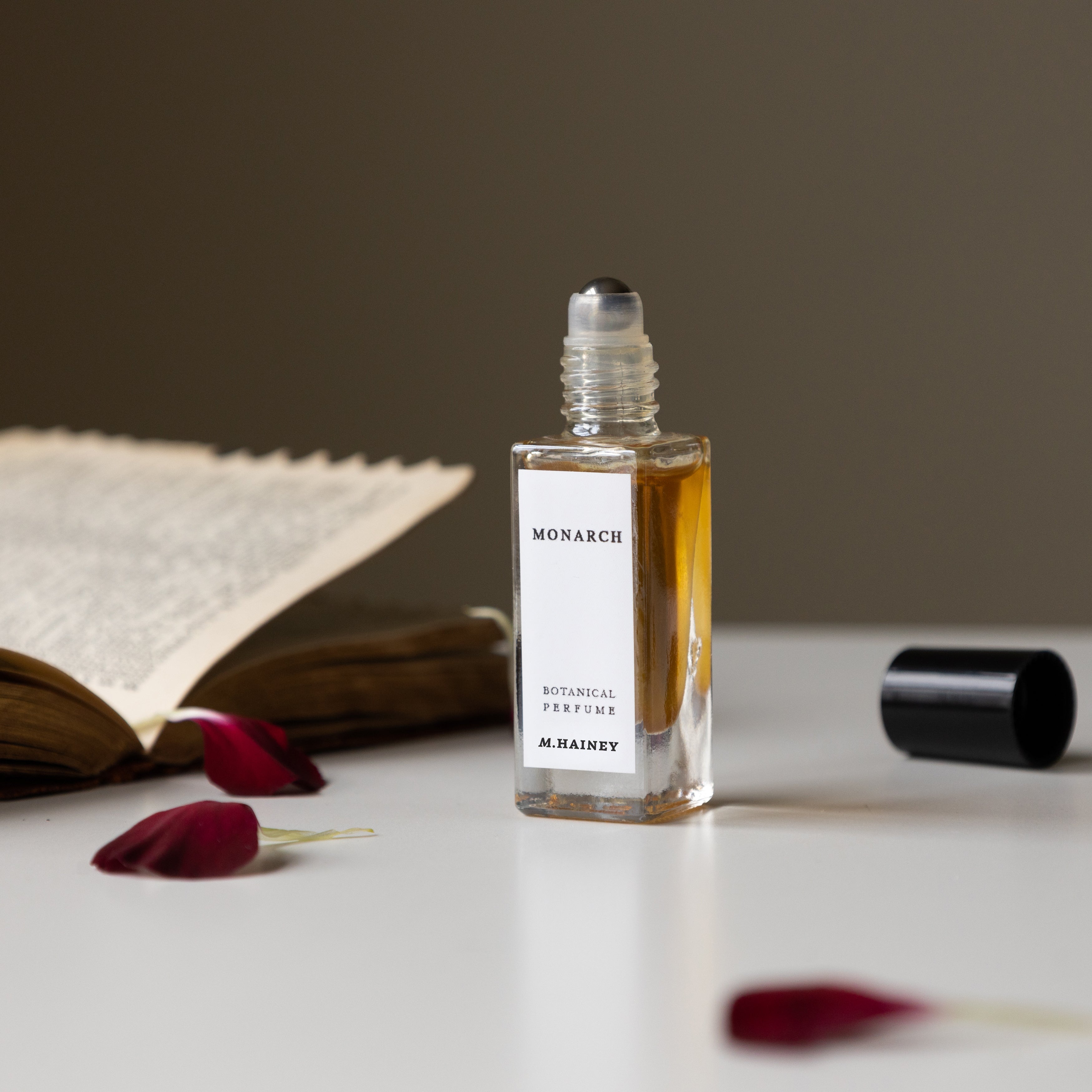 Roll-on perfume bottle labeled 'Monarch' by M. Hainey on a surface with red flowers and an open book.
