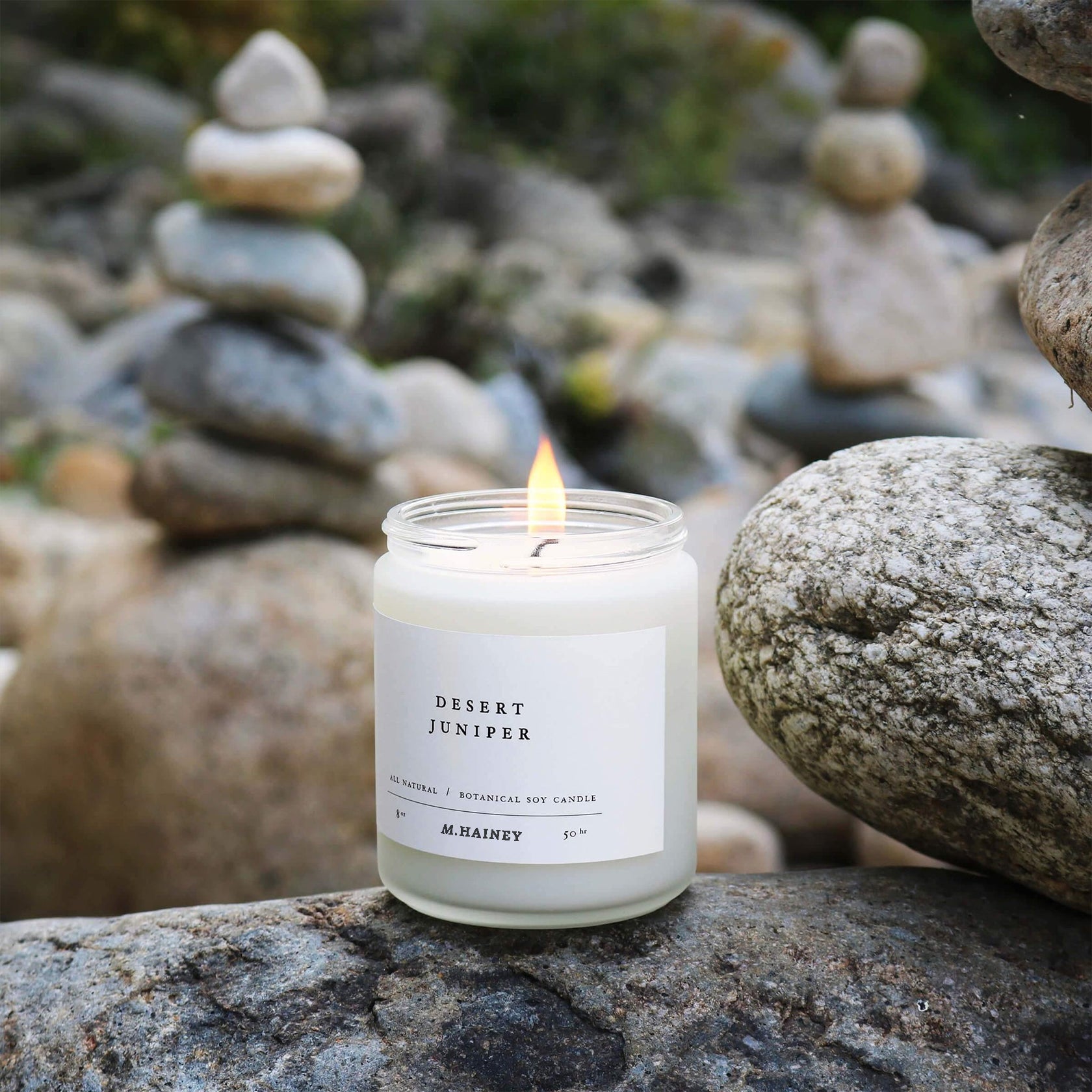 Candle labeled 'Desert Juniper' by M. Rainey on a rock with stone stacks in the background