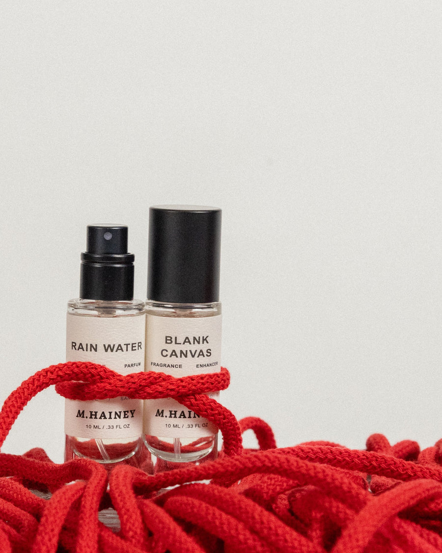 Two perfume bottles labeled 'Rain Water' and 'Blank Canvas' by M. Hainey on a red rope background.
